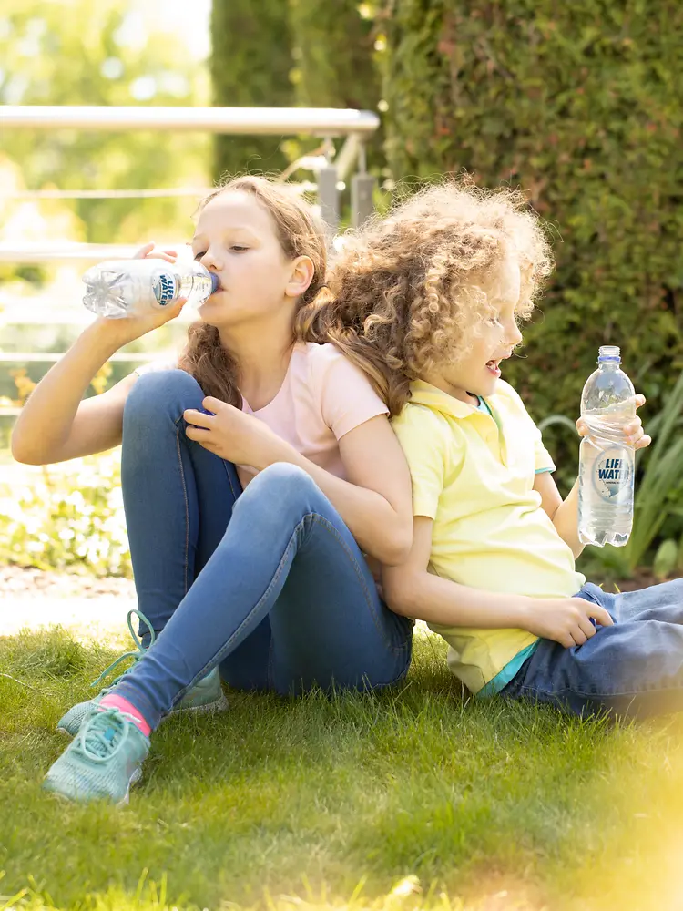 Dua anak sedang minum air kemasan di rumput.