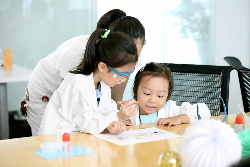 Dua anak dan seorang wanita memakai jas lab mewarnai gambar di meja