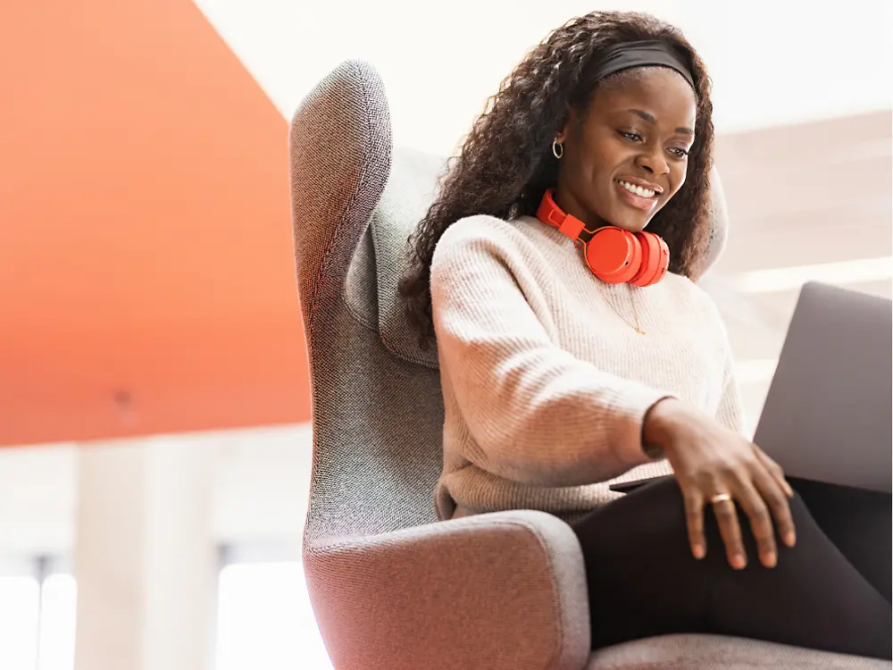 A Henkel employee sits in a comfy chair with her laptop on her knees. She smiles.