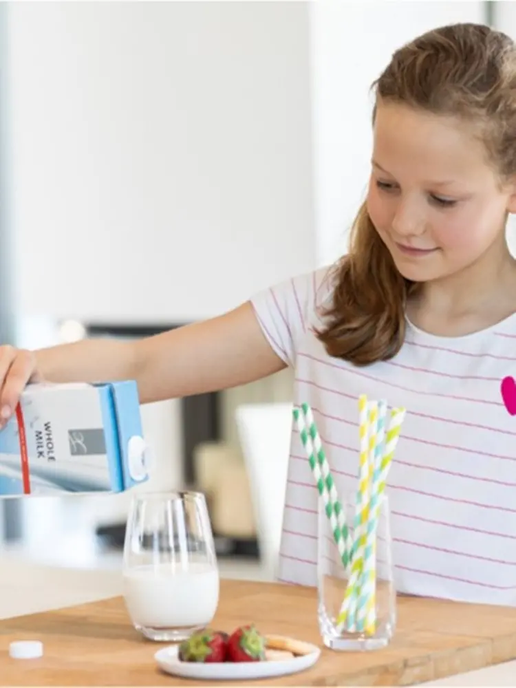 A girl pouring milk into a cup, with a glass of paper straws beside it.