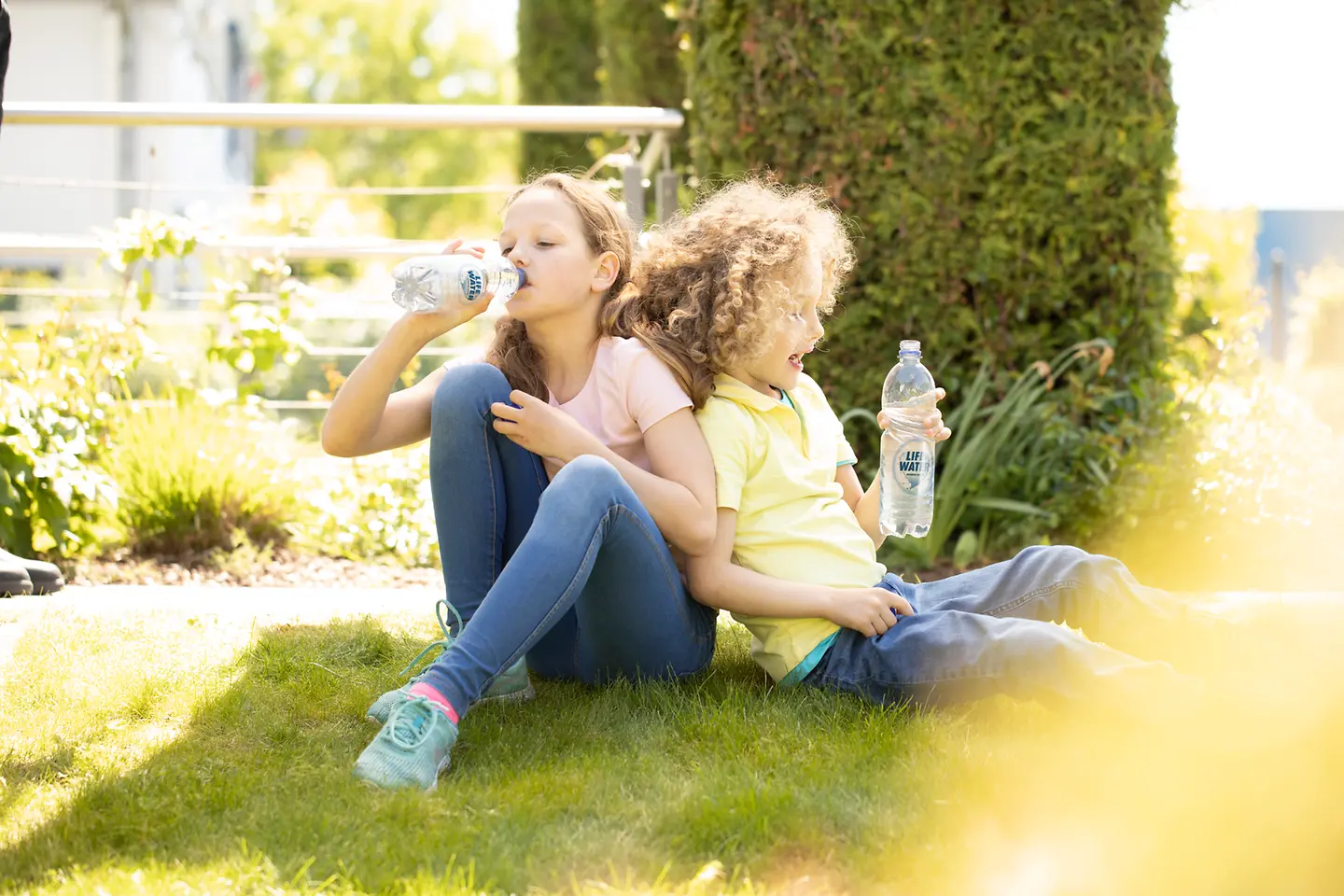 Two children drinking bottled water on the grass.
