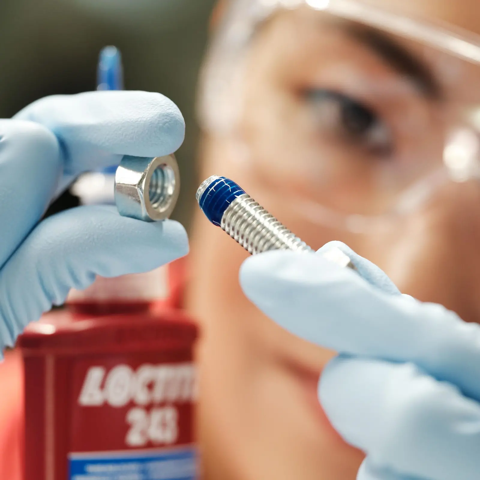 A woman's face in the background. In the front her hands with blue gloves holding a screw with blue Threadlocker.