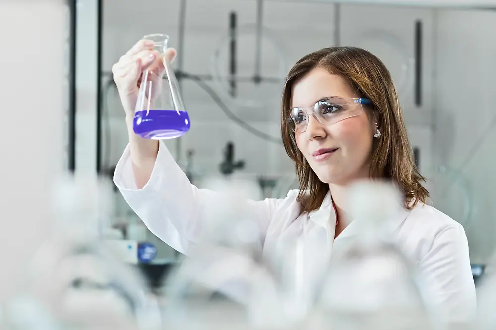 Seorang ilmuwan wanita sedang bereksperimen di laboratorium, mengenakan jas lab dan kacamata pengaman.