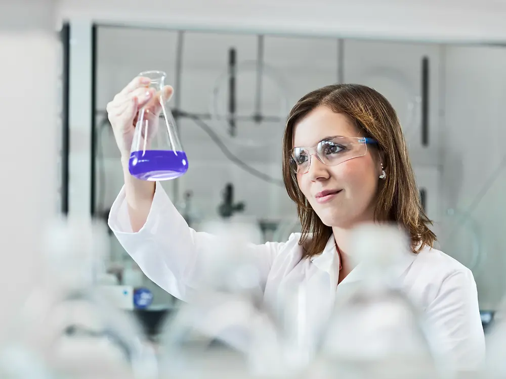 Seorang ilmuwan wanita sedang bereksperimen di laboratorium, mengenakan jas lab dan kacamata pengaman.
