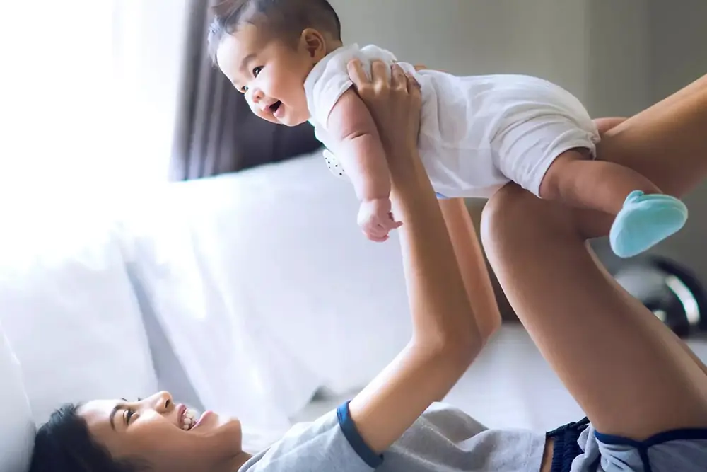 Ibu mengangkat dan bermain dengan bayi yang tersenyum di dalam rumah yang terang.