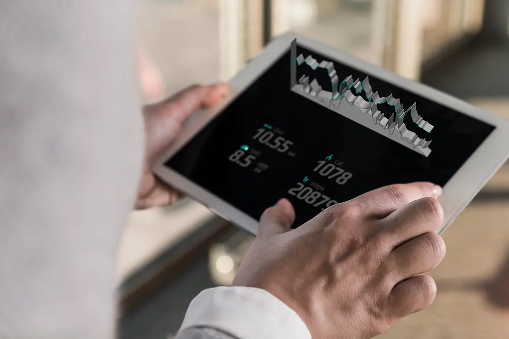 Seorang memegang tablet yang menampilkan grafik digital dan data numerik, dilihat dari belakang bahu dalam suasana ruangan yang terang.